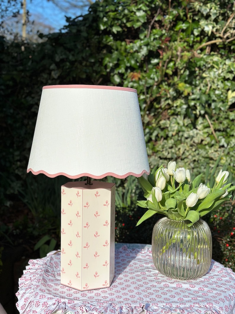 Op de afbeelding: Een witte lamp met een roze gegolfde rand op de lampenkap. De lampvoet is een witte achthoek met een roze bloemenpatroon. De lamp staat op een tafel met een roze bloemen tafelkleed.