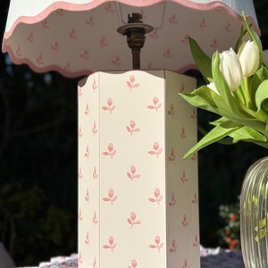 Op de afbeelding: Een witte lampenvoet met een roze bloemenpatroon en een witte lampenkap met een roze gegolfde rand. De lamp staat op een tafel met een bloemenpatroon.