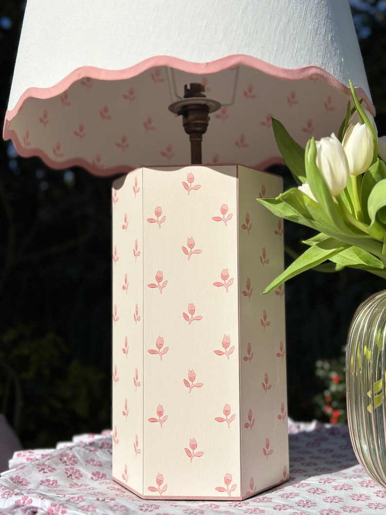 Op de afbeelding: Een witte lampenvoet met een roze bloemenpatroon en een witte lampenkap met een roze gegolfde rand. De lamp staat op een tafel met een bloemenpatroon.