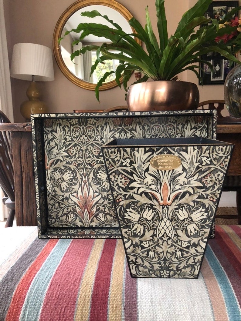 May include: A black and white tray and wastebasket with a floral pattern. The pattern features stylised flowers, leaves, and stems. The tray and wastebasket are made of wood and have a black finish. The wastebasket has a gold label with the text "William Morris"