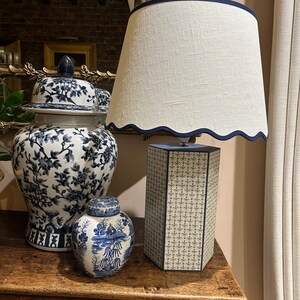 Op de afbeelding: Een witte lampenkap met een blauwe rand zit bovenop een blauwe en witte gepatroneerde lampenvoet. Twee blauwe en witte porseleinen gemberpotten staan op een houten tafel.