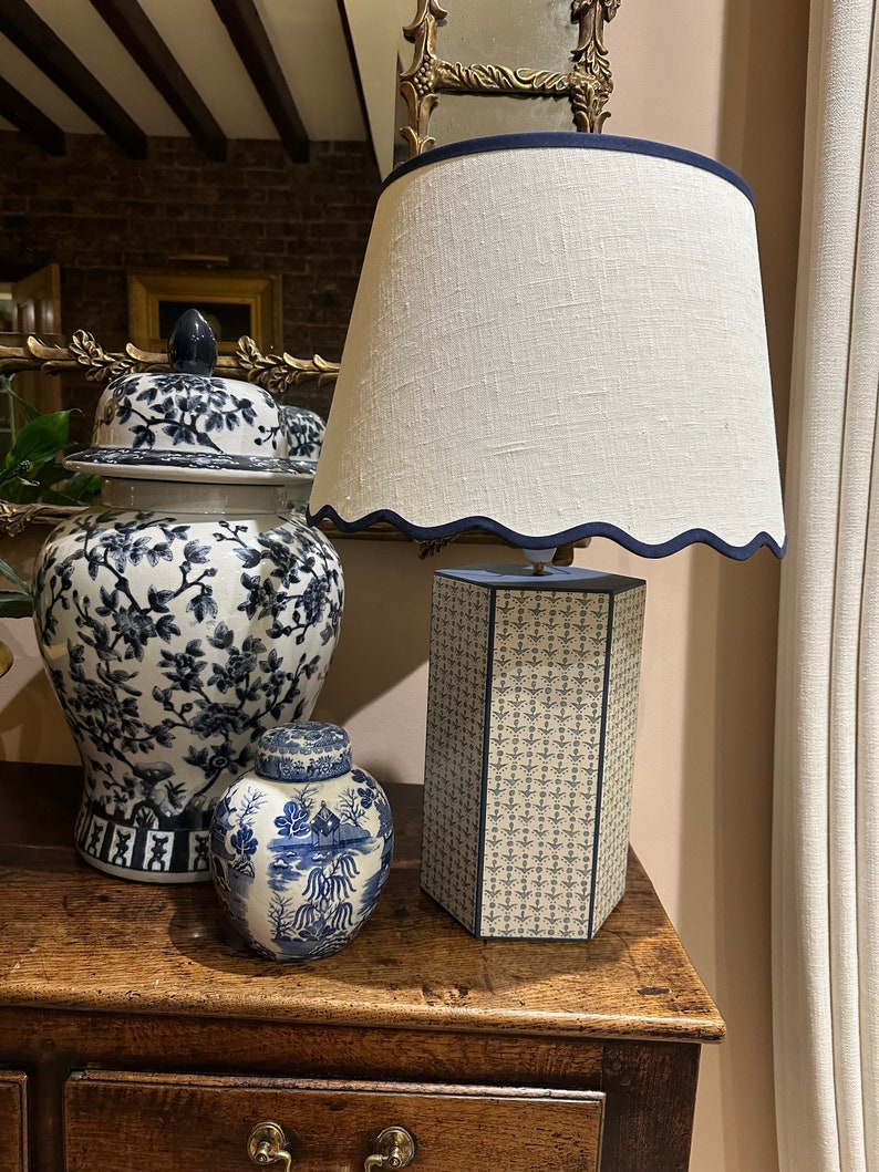 Op de afbeelding: Een witte lampenkap met een blauwe rand zit bovenop een blauwe en witte gepatroneerde lampenvoet. Twee blauwe en witte porseleinen gemberpotten staan op een houten tafel.