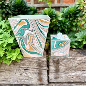May include: A green, gold and pink marbled wastebasket and tissue box set. The wastebasket is tall and has a square base. The tissue box is smaller and has a rectangular shape.