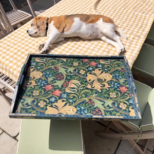 May include: A rectangular serving tray with a floral design in shades of blue, green, and gold. The tray has a dark blue border and handles. A dog is lying on a yellow and white checkered tablecloth.