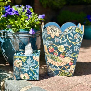 May include: A decorative tissue box and wastebasket set. The set features a dark teal background with a floral and bird design in shades of peach, green, and blue. The wastebasket has a scalloped top edge.