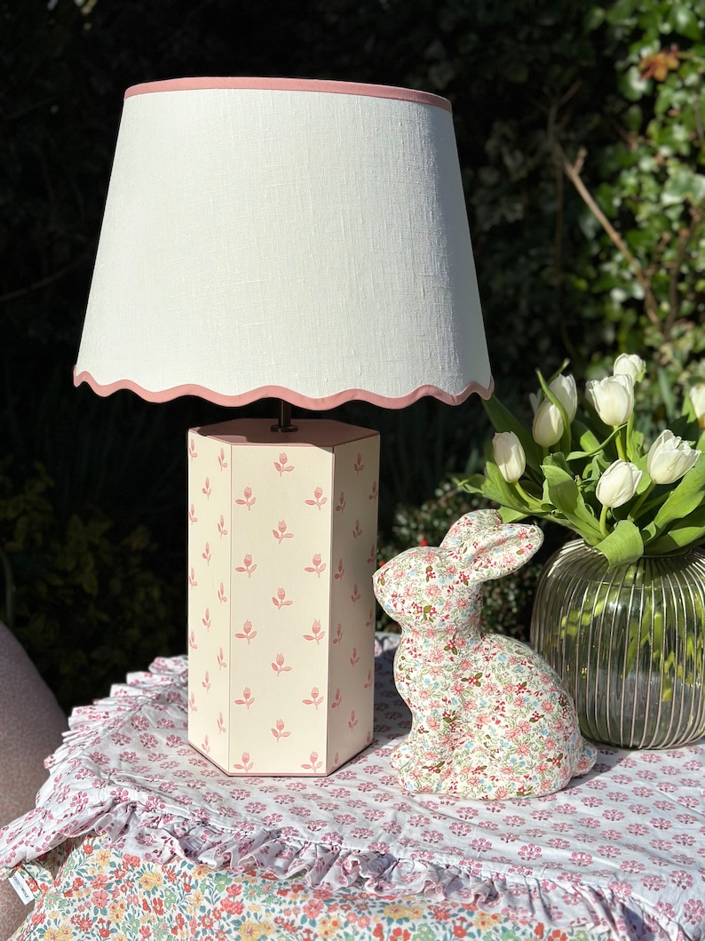 Op de afbeelding: Een witte lampenkap met een roze gegolfde rand zit bovenop een lampenvoet met een roze en wit bloemenpatroon. Een keramiek konijnenbeeldje met een bloemenpatroon staat op een tafelkleed met een bloemenpatroon.