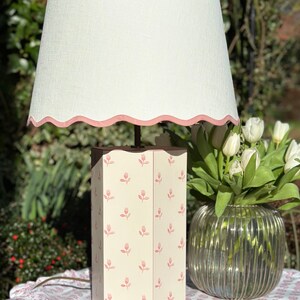 Op de afbeelding: Een witte lamp met een roze gegolfde lampenkap en een witte voet met een roze bloemenpatroon. De lamp staat op een tafel met een bloemenkleed.