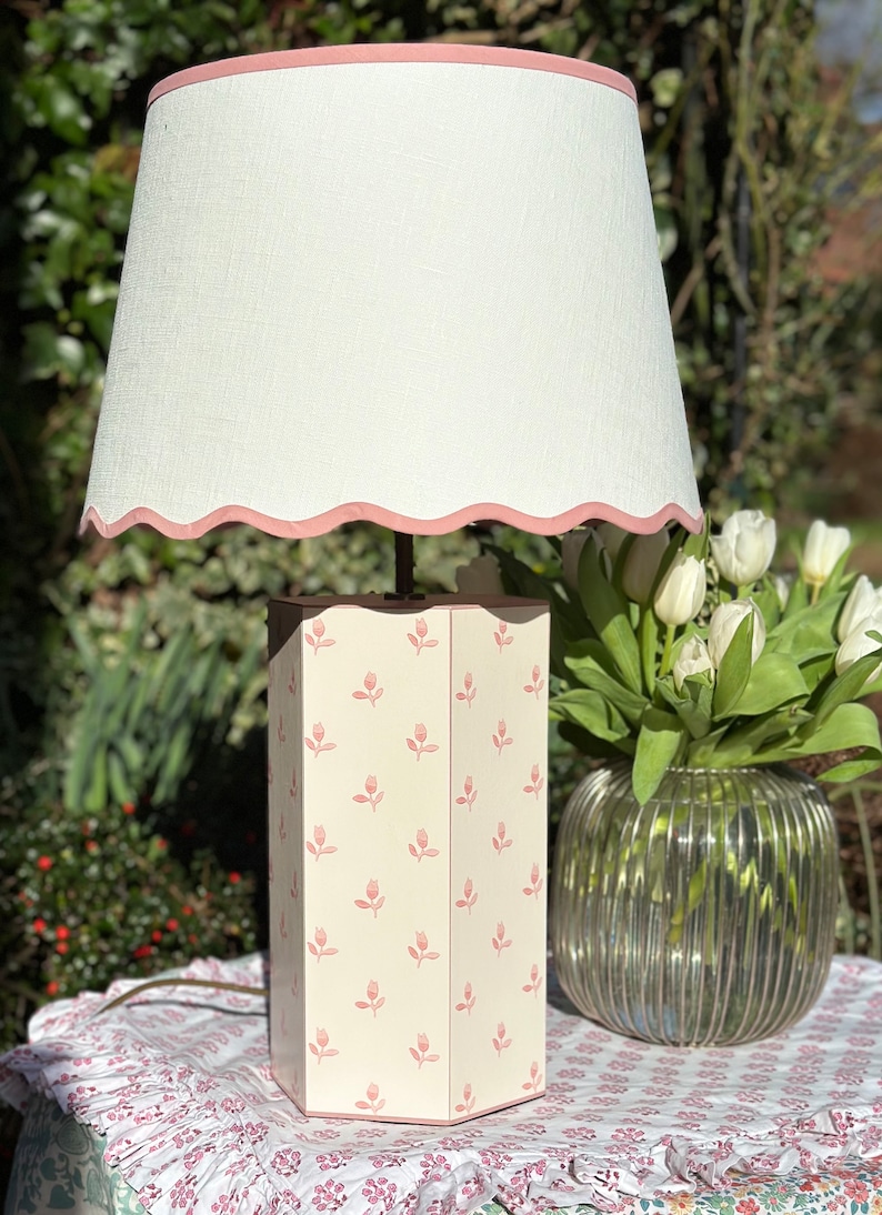 Op de afbeelding: Een witte lamp met een roze gegolfde lampenkap en een witte voet met een roze bloemenpatroon. De lamp staat op een tafel met een bloemenkleed.