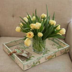 Puede incluir: Una bandeja con un diseño floral y un ramo de tulipanes amarillos en un jarrón de vidrio transparente. La bandeja tiene un borde verde y un fondo blanco con un patrón floral.