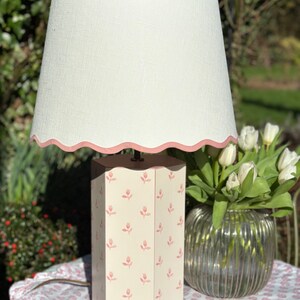 Op de afbeelding: Een witte lampenkap met een roze gegolfde rand zit bovenop een zeshoekige lampenvoet met een roze bloemenpatroon. De lamp staat op een tafel met een roze bloemenpatroon tafelkleed.