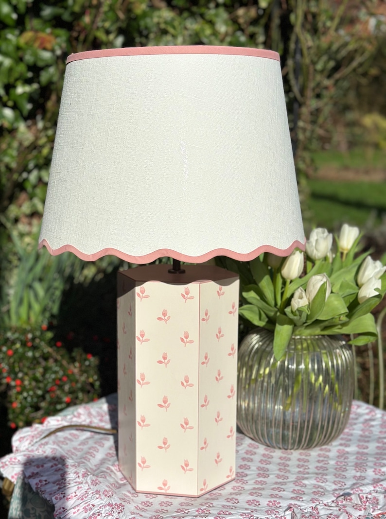 Op de afbeelding: Een witte lampenkap met een roze gegolfde rand zit bovenop een zeshoekige lampenvoet met een roze bloemenpatroon. De lamp staat op een tafel met een roze bloemenpatroon tafelkleed.