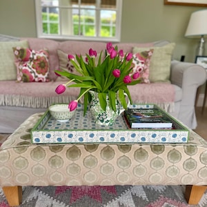 Könnte beinhalten: Ein dekoratives Ottoman mit einem Tablett, das eine Vase mit rosa Tulpen, eine kleine Schüssel und ein Buch trägt. Das Tablett hat ein blau-weißes Blumenmuster mit einem grünen Rand. Das Ottoman hat ein beige-grünes Muster, und der Teppich darunter hat ein rosa-graues geometrisches Design.