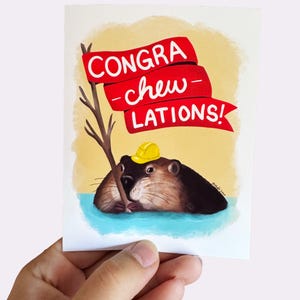 May include: A rectangular greeting card featuring a brown beaver wearing a yellow hard hat, holding a brown stick in blue water. Above the beaver is a red ribbon banner with the white text CONGRA-chew-LATIONS! set against a soft yellow background.