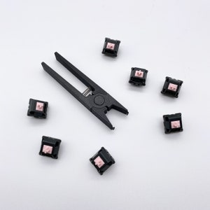 May include: A black plastic tool for removing mechanical keyboard keycaps, surrounded by eight black keycaps with pink accents.