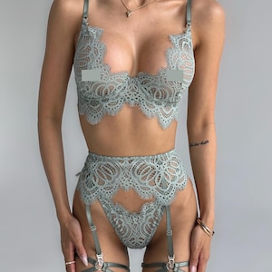 以下が含まれることがあります： セージグリーンのレースランジェリーセット。セットには、ブラジャー、ハイウエストブリーフ、調節可能なストラップ付きガーターベルトが含まれます。ブラジャーとブリーフは、複雑なフローラルレースのディテールが特徴です。ガーターベルトには金属リングのアクセントが付いています。ランジェリーは柔らかく繊細な生地で作られています。