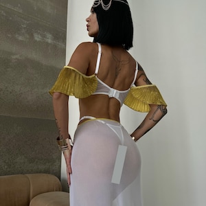 May include: A white sheer maxi skirt with a gold trim and a white lace bra with gold ruffled straps. The model is wearing a gold headpiece.