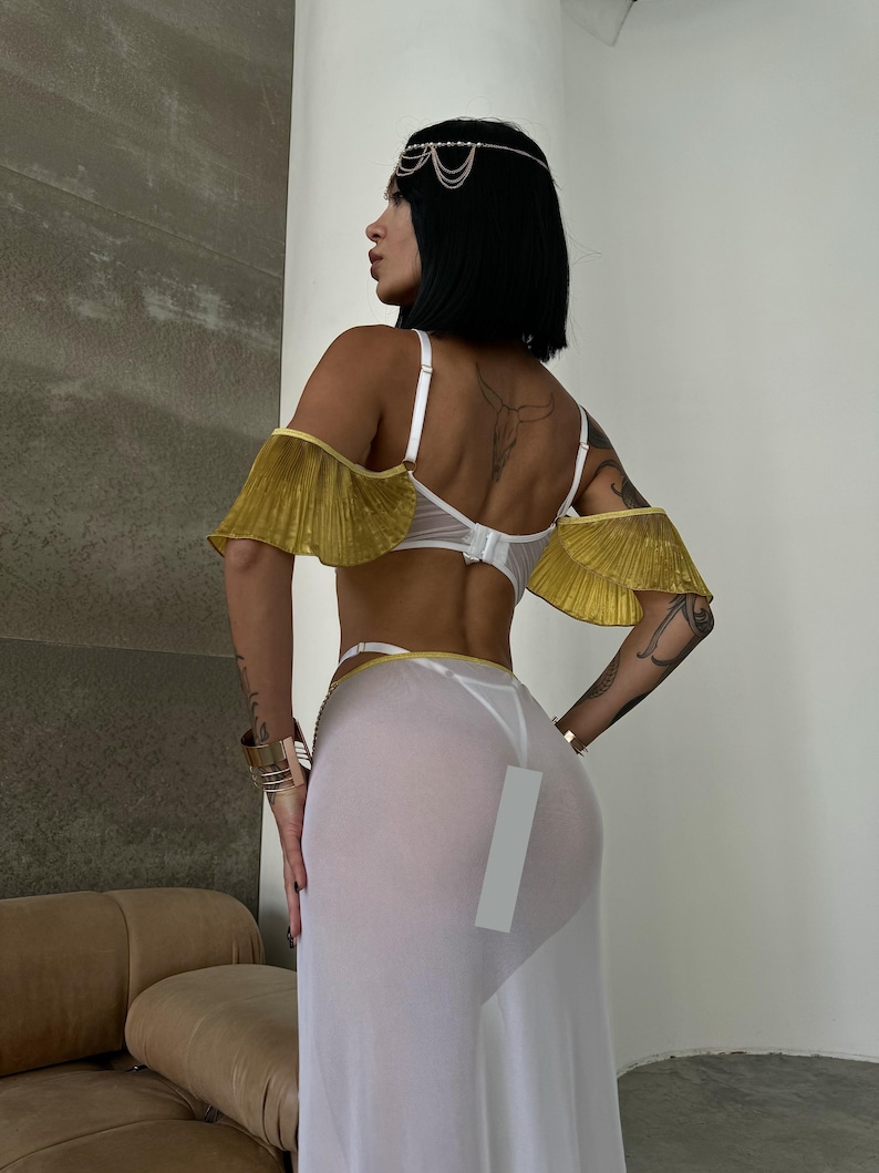 May include: A white sheer maxi skirt with a gold trim and a white lace bra with gold ruffled straps. The model is wearing a gold headpiece.