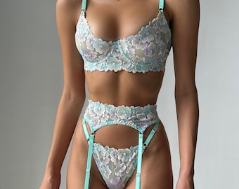 May include: A light blue lingerie set with floral lace detailing. The set includes a bra and garter belt with adjustable straps.