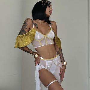 May include: White lingerie set with gold accents. The top features a mesh design with gold trim and a ruffled gold overlay on the shoulders. The bottom is a sheer skirt with a gold chain belt.