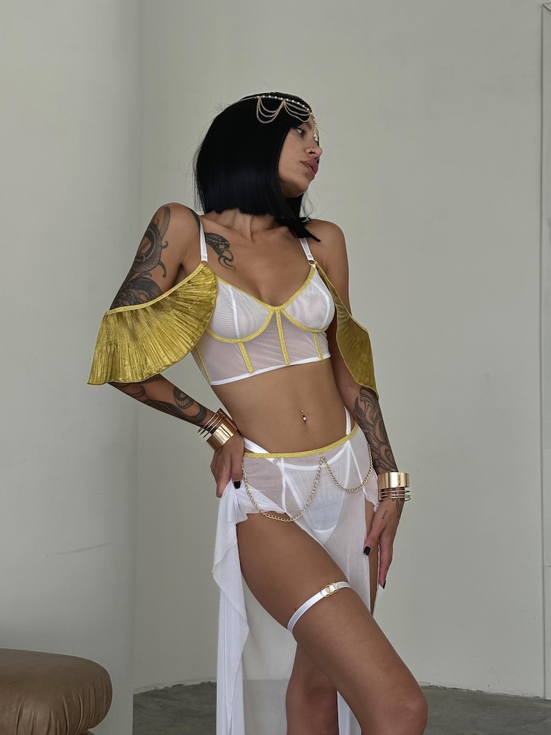 May include: White lingerie set with gold accents. The top features a mesh design with gold trim and a ruffled gold overlay on the shoulders. The bottom is a sheer skirt with a gold chain belt.