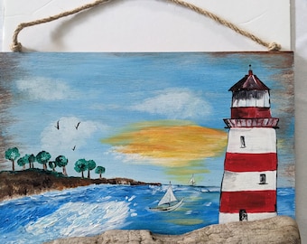 May include: A coastal-themed painting featuring a red and white striped lighthouse, sailboats, and a driftwood accent. The artwork depicts a seascape with blue water, a sunset, and a small island with green trees. A natural rope is attached for hanging.