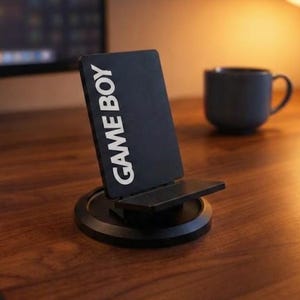 Peut inclure: Un support de téléphone rectangulaire noir avec le texte blanc GAME BOY imprimé verticalement sur la face avant. Le support possède une base circulaire et un petit rebord pour maintenir un appareil, fabriqué dans un matériau mat.