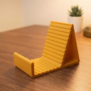 May include: A mustard yellow phone stand with a triangular profile and a ribbed, textured surface. The stand features a horizontal ledge at the base to support a mobile device, with a sloped backrest designed to hold a phone at an angle.