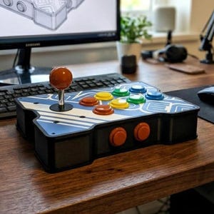 Puede incluir: Un mando arcade rectangular que cuenta con un joystick naranja y ocho botones de colores rojo, amarillo, verde y azul. La superficie superior muestra un patrón geométrico blanco y azul, mientras que en la parte frontal hay dos botones naranjas.