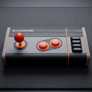 May include: A rectangular Commodore 64 game controller featuring a grey and black chassis with orange accents. It includes a red joystick, two circular red action buttons, and four black rectangular buttons labelled SELECT, START, LOAD, and SAVE.