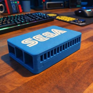 May include: A rectangular blue plastic case for a single-board computer featuring the white SEGA logo printed on the top surface. The case has a series of vertical ventilation slots along the side and multiple rectangular cutouts for ports.