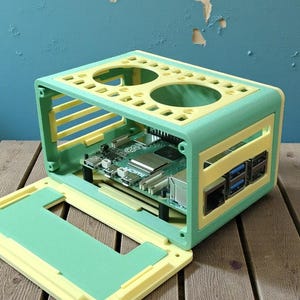 以下が含まれることがあります： ミントグリーンとイエローのツートンカラーのコンピューターケース。内部の回路基板が見えるオープンデザイン。ケースは角が丸い長方形で、上部に2つの大きな円形の開口部があります。フロントパネルは開いています。
