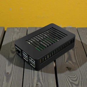 May include: A black rectangular electronic device with a slotted top and multiple ports on the side. The device is on a wooden surface with a yellow background. The device appears to be a computer component or enclosure.