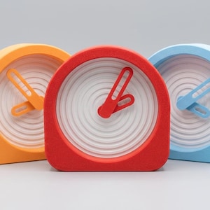 May include: Three analog clocks with rounded tops and matte finishes in orange, red, and light blue. Each clock features a white face with concentric circular ridges and matching colored hands with elongated slots in the center.