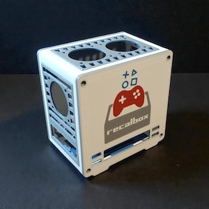 May include: A white and gray cube-shaped electronic device with the word "recalbox" and a red game controller logo. The device has a circular opening on the side and two circular openings on top.