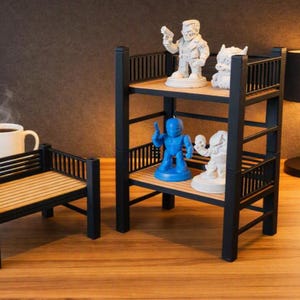 May include: A two-tier black display shelf with light wood slats and railings, holding four small character figurines. The top shelf features two white figures, while the bottom shelf displays one blue and one white figure. A separate low bench sits nearby.