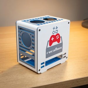 Puede incluir: Una caja de ordenador rectangular blanca y azul con un gráfico de mando de juego rojo y el texto recalbox en el lateral. El panel superior tiene dos aberturas circulares, y el panel lateral incluye una gran apertura circular y ranuras.