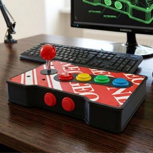 Puede incluir: Un mando arcade negro con un joystick de bola roja, cuatro botones de acción principales en rojo, amarillo, verde y azul, y dos botones laterales rojos. La superficie superior muestra un patrón de logotipo Neo Geo rojo y blanco con tornillos.