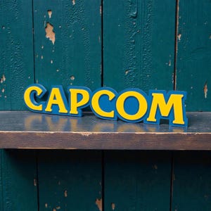 Könnte beinhalten: Ein blau-gelbes CAPCOM-Logo-Schild. Die Buchstaben sind gelb mit blauem Umriss und Sockel. Das Schild wird auf einem dunklen Holzregal vor einem teal-farbenen Holzgrund angezeigt.