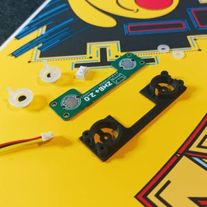 May include: An overhead shot displays electronic components on a yellow surface. A green circuit board with the text "ZMB+2.0" is next to a black plastic piece. White plastic and electrical connectors are also visible.