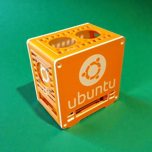 May include: An orange cube-shaped computer case with the Ubuntu logo and text. The case has a white trim and a fan on one side. The top has two circular openings. The case is on a green surface.