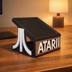 Puede incluir: Un soporte de exhibición negro inclinado con el logotipo blanco de Atari y el texto 2600 en el panel frontal. El soporte tiene un gráfico blanco estilizado en el lateral y una superficie superior plana e inclinada, con acabado mate.