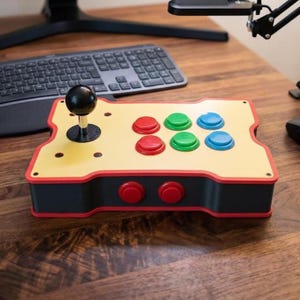 Puede incluir: Un mando arcade rectangular con un panel superior amarillo claro, un joystick negro y seis botones principales en rojo, verde y azul. El dispositivo tiene una base gris oscuro con borde rojo y dos botones rojos adicionales en la parte frontal.