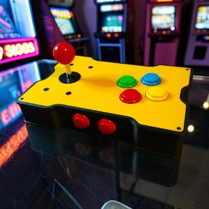 Pode incluir: Um comando arcade amarelo com base preta, apresentando um joystick de bola vermelha e quatro botões de ação principais em verde, azul, vermelho e amarelo. Dois botões vermelhos adicionais estão posicionados na parte frontal da caixa preta.