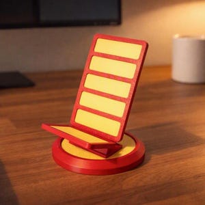 May include: A red and yellow phone stand featuring a circular base and an upright rectangular support with five horizontal slots. The structure is constructed with a textured finish and provides a stable platform for holding a mobile device.