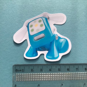 Puede incluir: Una pegatina de robot perro azul con un contorno blanco. El robot perro tiene una cara azul claro con un patr&oacute;n de luz amarillo y verde. La pegatina mide aproximadamente 6 cm de largo.