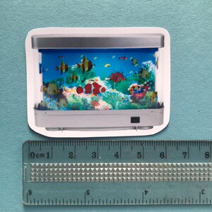 Puede incluir: Una pegatina de un peque&ntilde;o acuario rectangular plateado con un fondo azul y peces coloridos nadando dentro. El acuario tiene un interruptor de luz en la parte inferior derecha.