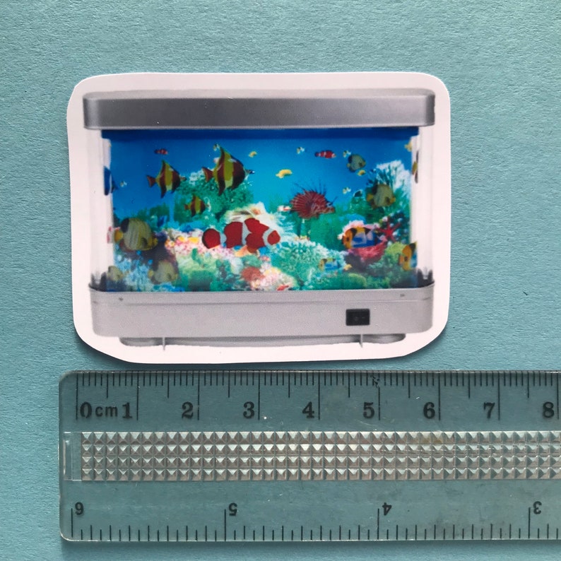 Puede incluir: Una pegatina de un peque&ntilde;o acuario rectangular plateado con un fondo azul y peces coloridos nadando dentro. El acuario tiene un interruptor de luz en la parte inferior derecha.