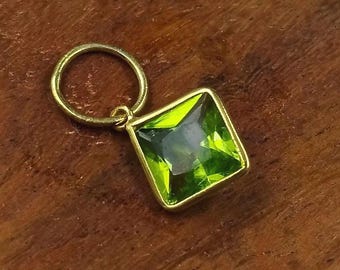 May include: A gold pendant featuring a square, vivid green gemstone. The gemstone is set within a square frame and connected to a small circular loop. The pendant is shown on a brown wooden surface.