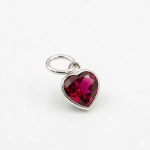 Pode incluir: Um pingente em forma de coração com uma pedra preciosa vermelha escura facetada, engastada em metal prateado. O pingente tem uma pequena argola na parte superior para prender a uma corrente ou pulseira. A pedra preciosa tem uma cor vibrante e rica.