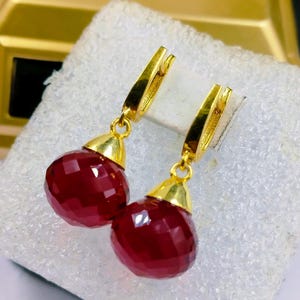 May include: A pair of gold earrings featuring faceted, deep red teardrop pendants. The earrings have a lever-back closure. The pendants are a rich, dark red colour and catch the light, creating a sparkling effect. The earrings are displayed on a white surface.